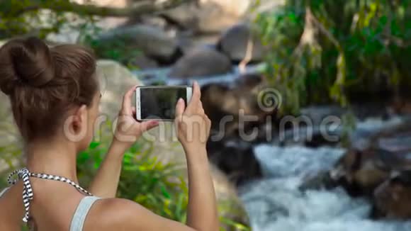 年轻女子在丛林森林中的山瀑布中用手机做照片景观女游客视频的预览图