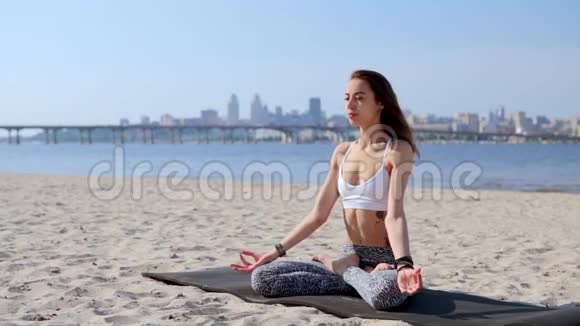 一位美丽放松的年轻女子坐在海滩上的莲花姿势的肖像有着海洋和城市的背景妇女视频的预览图