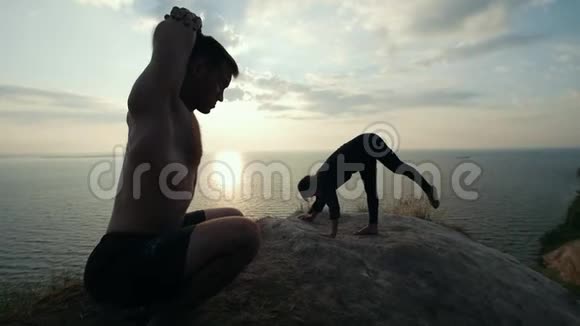 日出时在悬崖上练习瑜伽的年轻夫妇剪影视频的预览图