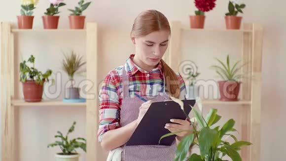 小生意开一家花店一个穿着格子衬衫和条纹围裙的女孩拿着文件夹写着视频的预览图