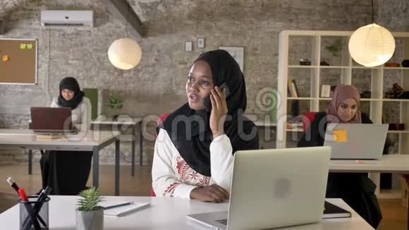 戴着头巾的年轻黑人穆斯林妇女在现代办公室里打电话三个漂亮的穆斯林妇女坐在那里工作视频的预览图