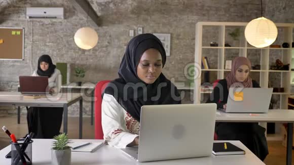 年轻的穆斯林黑人女士戴着头巾在笔记本电脑上打字在镜头中微笑三位美丽的穆斯林妇女在视频的预览图