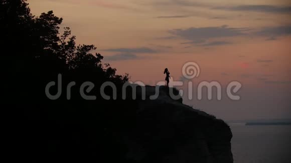 日落时分年轻女子在河边悬崖上跳舞的剪影视频的预览图
