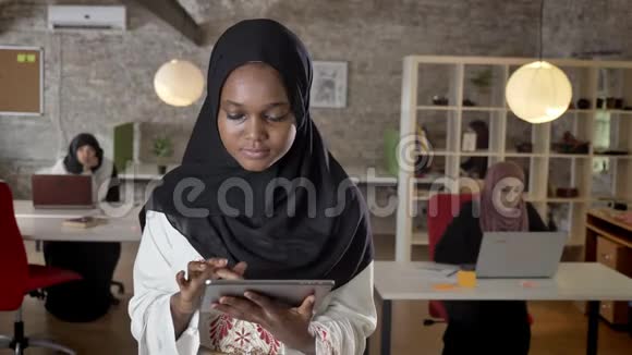 年轻的穆斯林黑人妇女戴着头巾使用平板电脑看着镜头微笑着伊斯兰商业妇女在笔记本电脑上打字视频的预览图