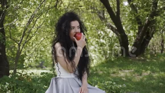 美丽的黑发姑娘坐在夏日花园里吃红苹果视频的预览图