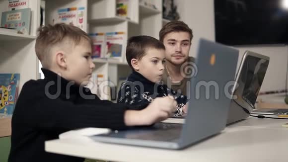 年轻迷人的男老师教两个小男孩如何使用笔记本电脑等小工具技术和进步视频的预览图