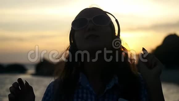 戴着耳机的年轻女子的剪影在美丽的夕阳下在海滩上欢快地跳舞听音乐视频的预览图