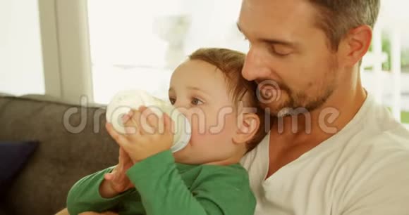 男孩在父亲的膝上在家沙发上喝牛奶视频的预览图