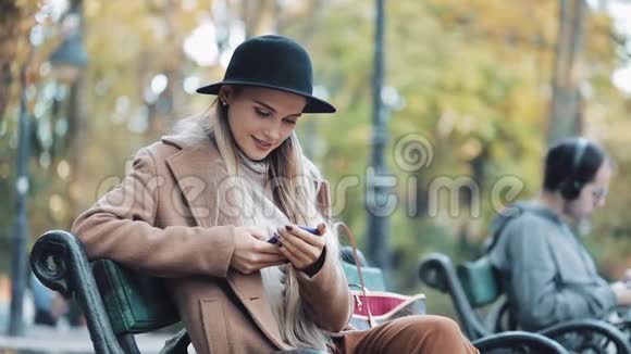 穿着智能手机的漂亮女人在秋天公园的长凳上放松户外技术视频的预览图