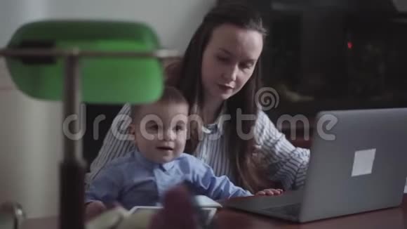 母亲和小儿子坐在室内桌子上的笔记本电脑的肖像忙碌的年轻母亲在家工作孩子视频的预览图