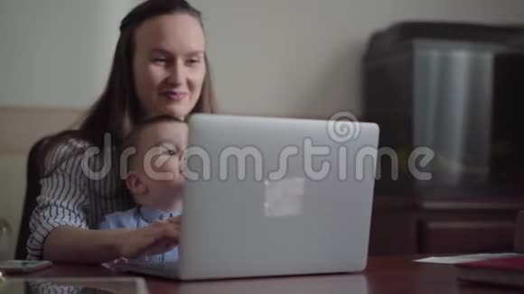 肖像母亲和小儿子坐在室内的桌子上拿着笔记本电脑忙碌的年轻母亲在家工作孩子视频的预览图