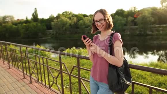 戴眼镜的微笑女孩用智能手机站在桥上的户外视频的预览图