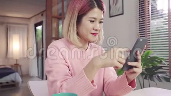年轻的亚洲女人一边使用智能手机一边躺在客厅的桌子上快乐女用手机发短信视频的预览图