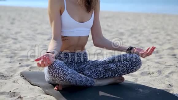 一位美丽放松的年轻女子坐在海滩上的莲花姿势的肖像有着海洋和城市的背景妇女视频的预览图