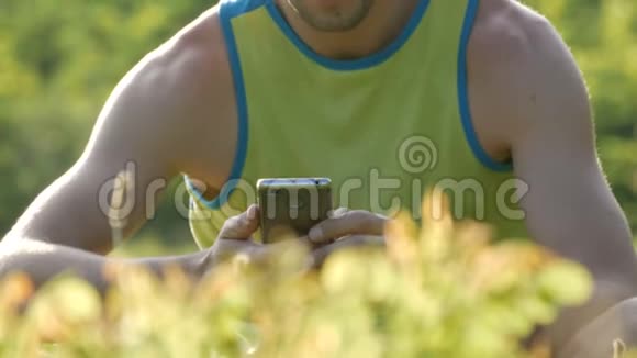 夏天在美丽的大自然背景下一个年轻可爱的男人在绿草上看智能手机视频的预览图