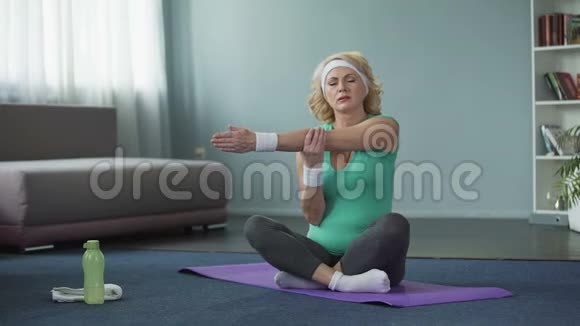 身穿运动服坐在瑜伽垫上按摩肩膀的金发老年妇女视频的预览图