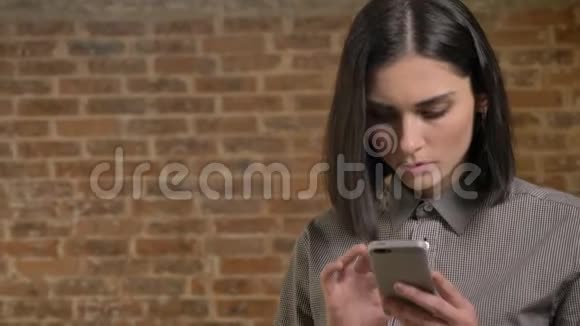 年轻漂亮的女人在电话上打字咬嘴唇的画像集中砖墙背景视频的预览图