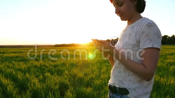 一位年轻迷人的女士正在一片绿色草坪上用一部智能手机在夕阳的背景下缓慢移动视频的预览图
