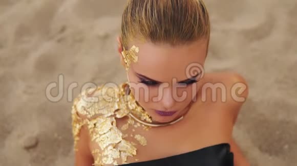 特写年轻模特用专业的金色艺术化妆躺在海滩上看着镜头吸引力视频的预览图