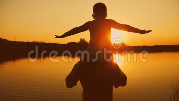 父亲和儿子在海滩上玩耍的剪影男孩举起双手在美妙的日落中模仿飞行视频的预览图