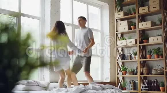 年轻的已婚夫妇正在双人床上跳着舞房间里有大窗户快乐的人们玩得很开心视频的预览图
