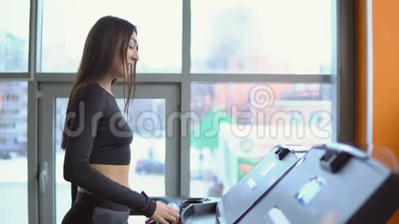 一位年轻的黑发女子在健身房的跑步机上站在窗口背景下视频的预览图