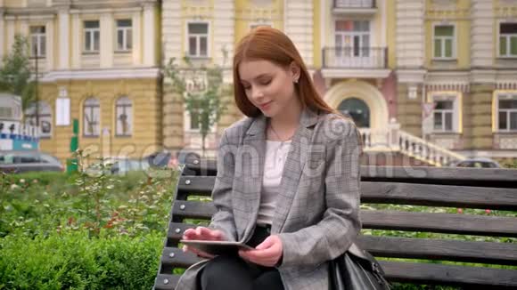 年轻漂亮的姜女穿着夹克坐在长凳上拿着石碑城市街道背景视频的预览图