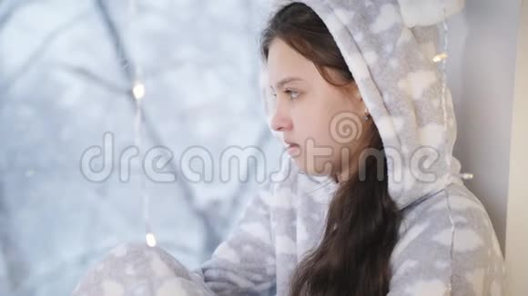 少女温特穿着暖和的睡衣和围巾坐在窗边看起来窗外正在下雪视频的预览图