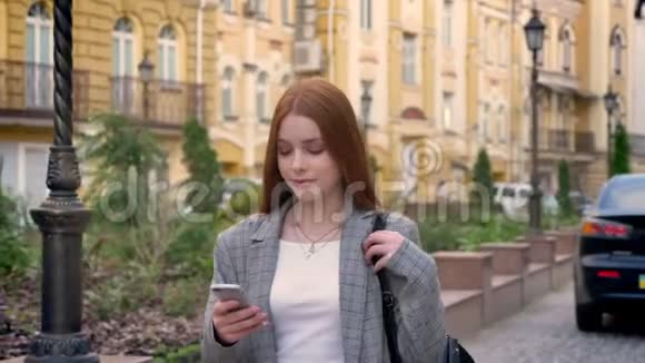 年轻漂亮的姜女走在街上看着她的手机黄色的建筑背景视频的预览图