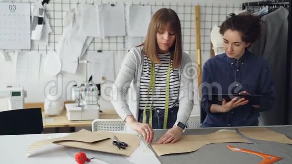 女服装设计师正在用粉笔在织物上绘制新服装的轮廓而她的同事正在帮助她和视频的预览图