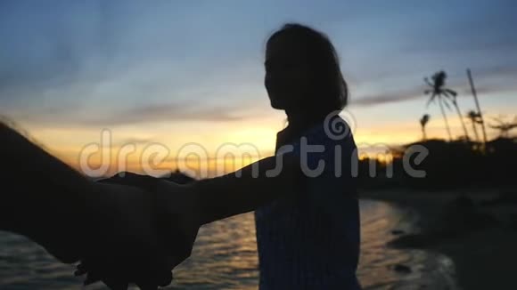 年轻女子的剪影牵着她朋友的手在美丽的夕阳下示意他沿着海滩散步来吧视频的预览图