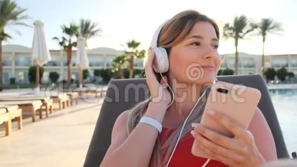 穿着红色比基尼带着白色耳机听智能手机音乐的年轻迷人的微笑少女的肖像视频的预览图