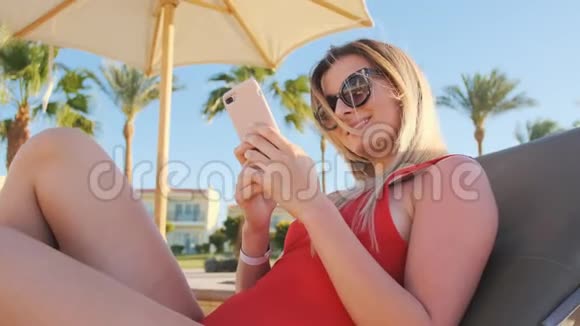 穿红色比基尼的女孩在度假时使用她的智能手机在酒店泳池边区域度假旅行妇女在水疗中心放松视频的预览图