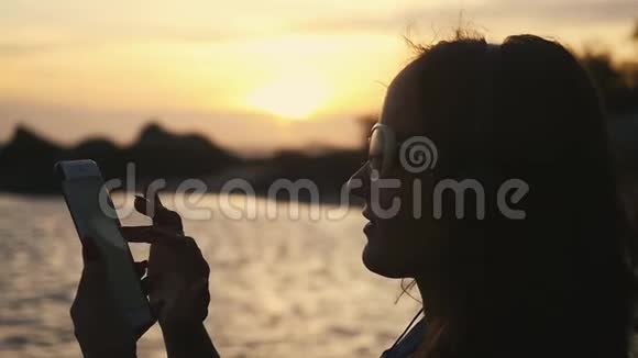 戴墨镜的年轻女性在热带海滩夕阳下使用手机的剪影有度假时间慢慢慢慢视频的预览图