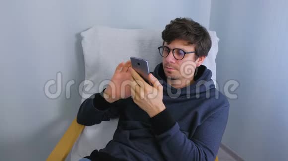 戴眼镜的男人坐在扶手椅上用智能手机浏览社交媒体视频的预览图