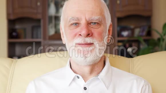 穿着白色马球衫坐在家里沙发上的英俊微笑老人的肖像视频的预览图