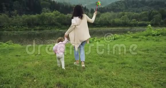 母亲带着女儿跑到河边牵着手母亲的快乐时刻山脉背景风车视频的预览图