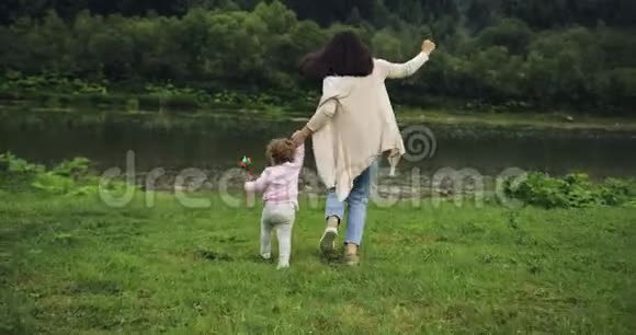 母亲带着女儿跑到河边牵着手母亲的快乐时刻山脉背景风车视频的预览图