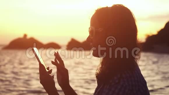 年轻漂亮的女士戴着太阳镜在热带海滩惊人的日落下使用手机慢动作1920x1080视频的预览图