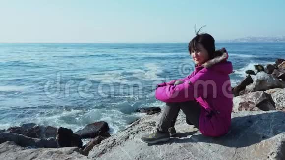 白种人年轻的女人穿着粉红色的外套坐在波浪海附近的岩石上视频的预览图
