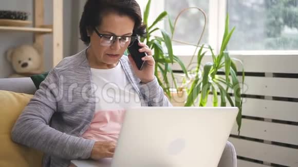 成熟的女商人正在打电话在家庭内部使用笔记本电脑视频的预览图