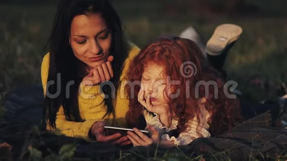 美丽的母亲和她可爱的小女儿躺在公园的草地上在智能手机上看视频视频的预览图