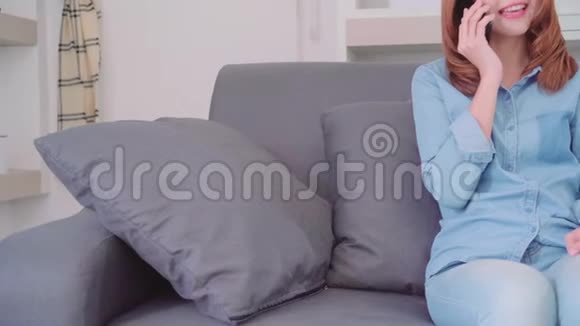 亚洲女人在家里的客厅里放松时一边躺在沙发上与朋友用智能手机交谈视频的预览图