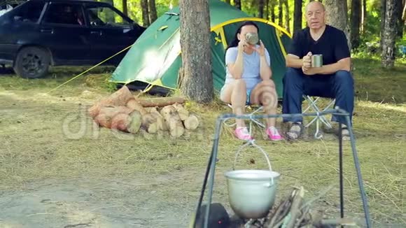 游客们一男一女坐在帐篷前喝着茶看着挂在火堆上的保龄球视频的预览图