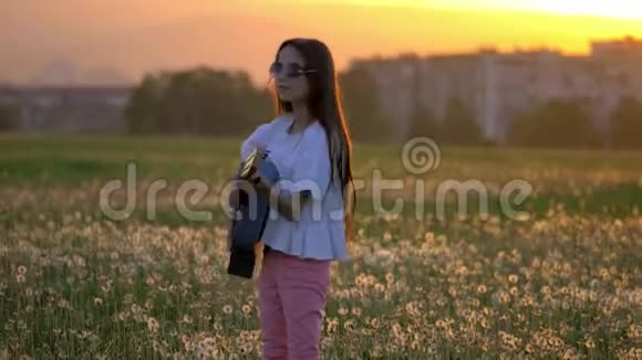 日落时美丽的小女孩在蒲公英地弹吉他远离城市建筑的户外情感和乐趣视频的预览图