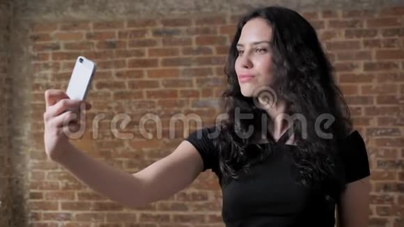 年轻漂亮的黑发女孩正在智能手机上自拍摆姿势交流理念砖背景视频的预览图