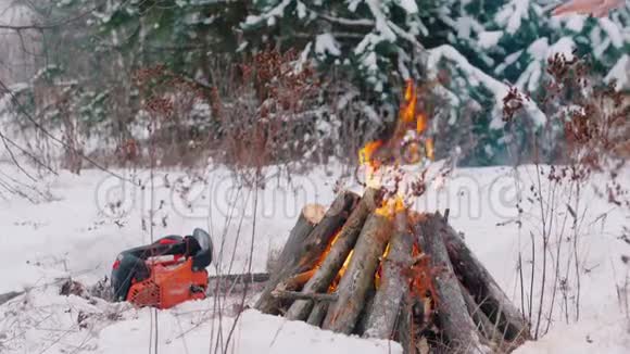 篝火晚会点燃了冬林中的火种一个人坐在火旁温暖他们的手一只链锯躺在地上视频的预览图