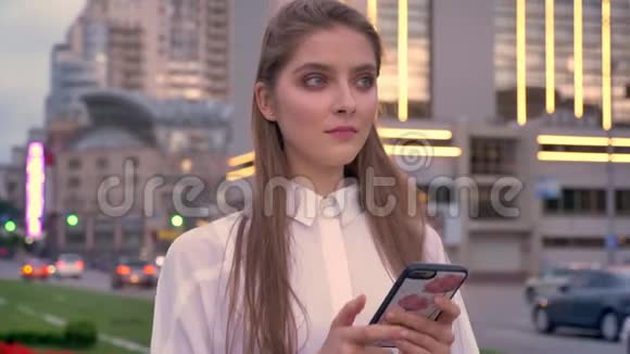 一个年轻漂亮的女孩在夏天的城市中心的日落时分看着她的智能手机视频的预览图