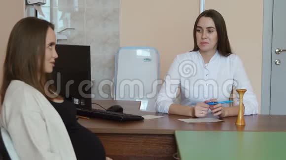 年轻的孕妇在医生的接待穿着白色的晨衣坐在桌边的医生问病人视频的预览图