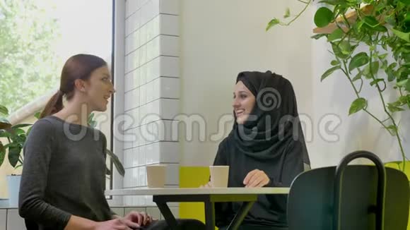 两个年轻漂亮的女人坐在咖啡馆里聊天其中一个是戴着头巾的漂亮穆斯林女人微笑着视频的预览图